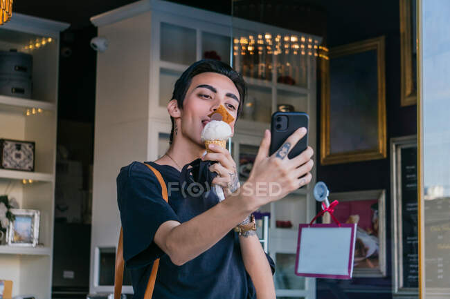 Content gay male eating sweet ice cream and taking self shot on mobile phone in city — Stock Photo