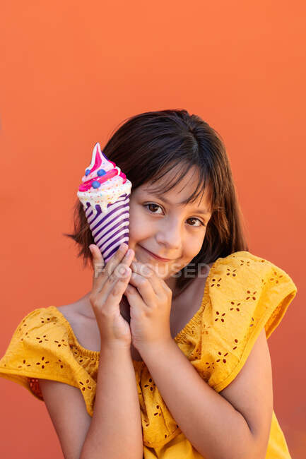 Aufrichtiges Kind in gelber Kleidung mit braunen Augen und leckerem Eis, das in die Kamera auf orangefarbenem Hintergrund blickt — Stockfoto