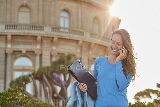 Щаслива доросла жінка в повсякденному вбранні говорить на смартфоні з папкою в руці, стоячи біля зелених рослин і старої будівлі в парку в сонячний день — стокове фото