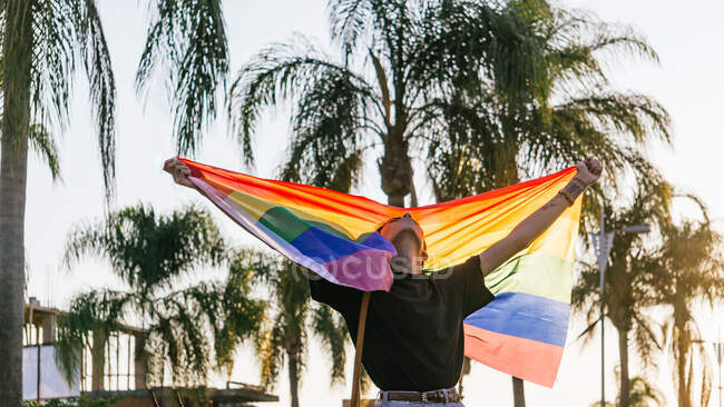 Delighted male gay standing with eyes closed raising rainbow LGBT flag during sunset in city — Stock Photo