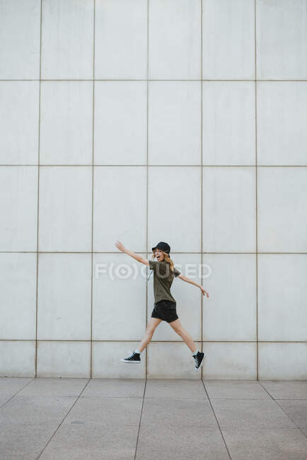 Vista lateral de la alegre hembra en ropa casual y zapatos de goma saltando mirando a la cámara con los brazos extendidos por encima de la pasarela de azulejos en la ciudad - foto de stock