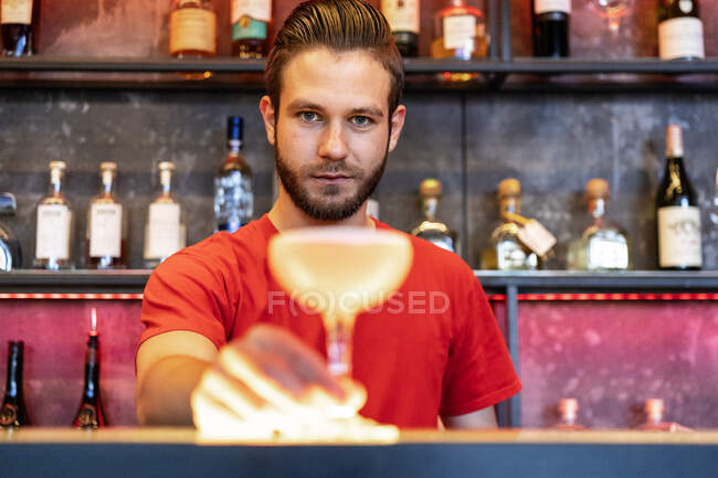 Веселий бармен подає склянку з алкогольним коктейлем на лічильнику в барі і дивиться на камеру — стокове фото