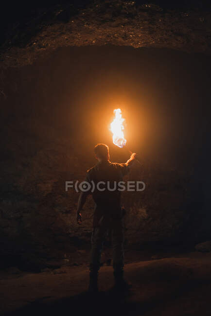 Back view of unrecognizable male speleologist with flaming torch standing in dark narrow rocky cave while exploring subterranean environment — Stock Photo
