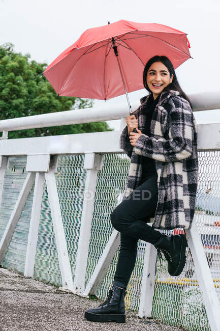 Vista lateral de una mujer étnica optimista con paraguas sonriendo y mirando hacia otro lado mientras se apoya en la barandilla del puente en un día lluvioso en el parque - foto de stock