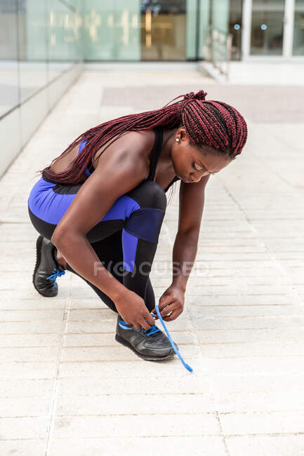 Mujer atlética étnica sentada en la calle y apretando cordones de zapatillas - foto de stock