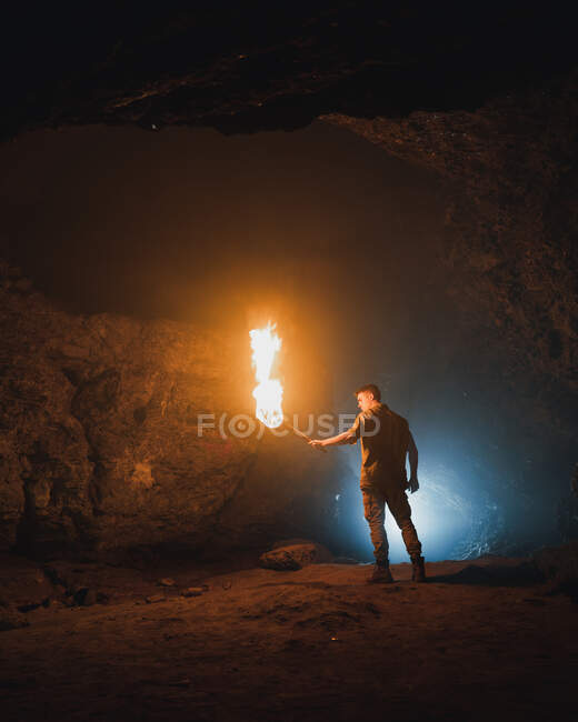 Back view of young male speleologist with flaming torch standing in dark narrow rocky cave while exploring subterranean environment — Stock Photo