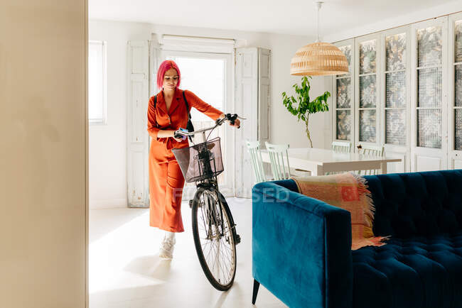 Young creative female designer in trendy outfit and eyeglasses using smartphone while standing with bicycle in modern light apartment — Stock Photo