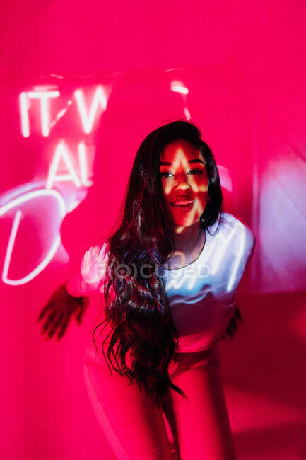 Glückliche junge hispanische Frau blickt in die Kamera, während sie sich an die Wand in der Nähe von Neon lehnt — Stockfoto