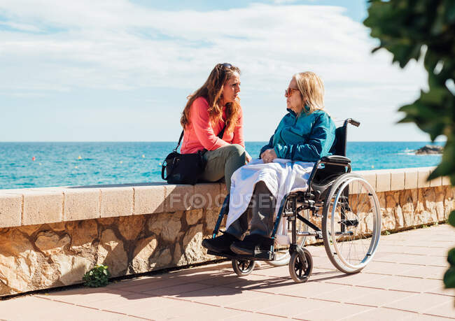 Adult woman and aged mother in wheelchair chilling on embankment and talking to each other on sunny day near sea — Stock Photo