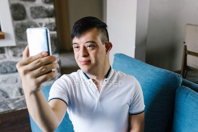Sorrindo adolescente latino menino com síndrome de Down tomando auto-tiro no smartphone enquanto sentado no sofá em casa — Fotografia de Stock