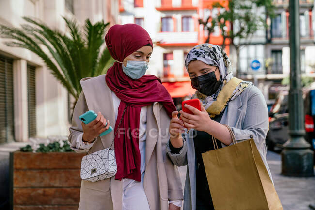 Mujeres amigas musulmanas en hiyabs y con bolsas de papel usando smartphone en la calle después de ir de compras - foto de stock