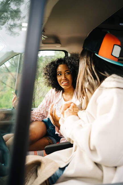 A través de la ventana de la joven mujer asiática alegre sentado en el asiento del conductor del vehículo de autocaravana mientras viaja con su novia a través de la naturaleza - foto de stock