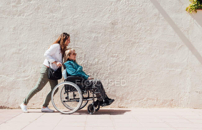 Side view of adult daughter pushing wheelchair with senior mother while walking along city street on sunny day — Stock Photo
