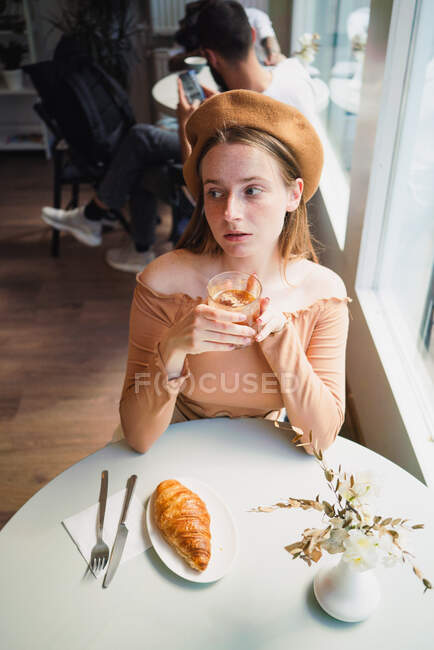 Hembra francesa en boina sentada a la mesa en la cafetería con vaso aromático de café y croissant recién horneado - foto de stock