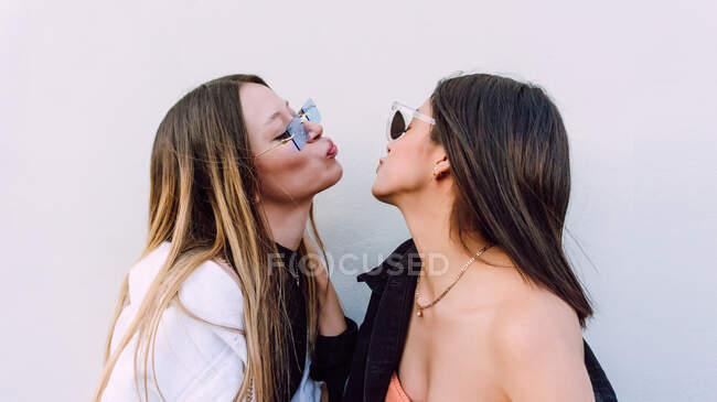 Вид сбоку подруг в стильных солнцезащитных очках с длинными волосами, делающих воздушные поцелуи друг с другом — стоковое фото