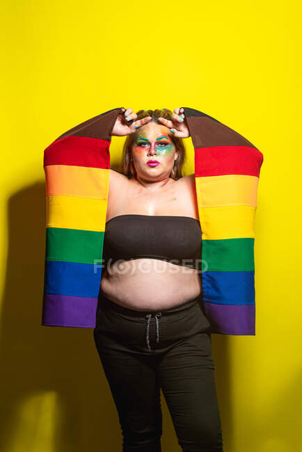 Modèle féminin en surpoids avec maquillage créatif montrant le drapeau LGBT et regardant la caméra sur fond jaune — Photo de stock