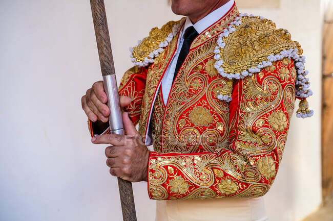 Crop anonymous picador with wooden spear and in bright red costume decorated with golden embroidery preparing for corrida festival — Stock Photo