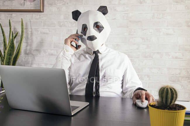 Empreendedor masculino irreconhecível em panda urso máscara falando no celular contra netbook durante teletrabalho na mesa — Fotografia de Stock
