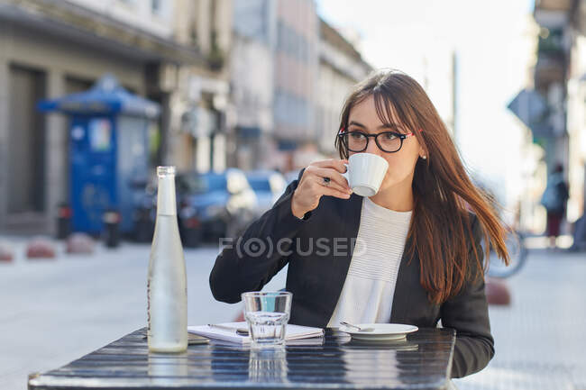 Modern young female entrepreneur in stylish outfit and glasses drinking coffee while sitting at table with notebook and bottle of water in urban outdoor cafeteria — Stock Photo