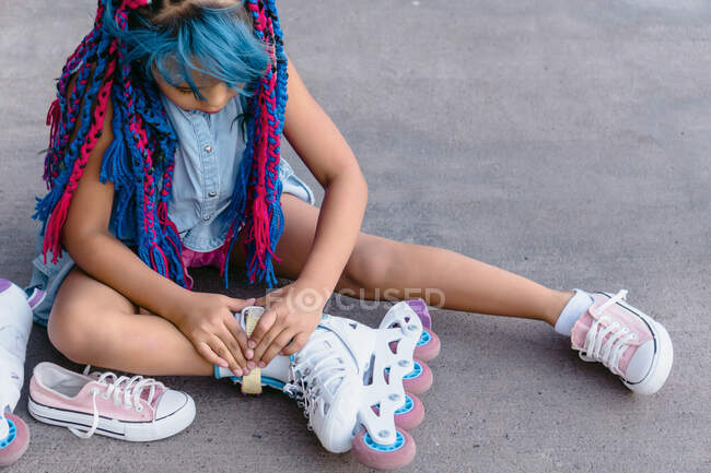 Crop ethnic kid with colorful braids putting on roller skate while sitting on walkway in daytime — Stock Photo