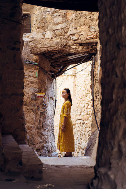 Vue latérale de la jeune voyageuse en robe jaune debout dans l'embrasure de la porte de la maison en pierre ruinée antique dans le vieux village Al Hamra à Oman — Photo de stock