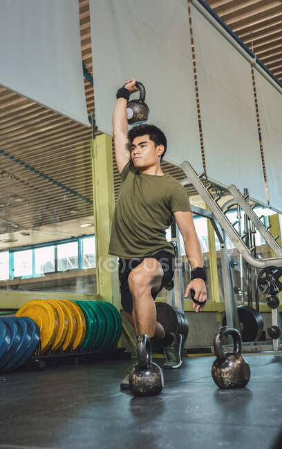 Homem asiático treinando ombros e braços com kettlebells pesados no ginásio durante o treino funcional e olhando para longe — Fotografia de Stock