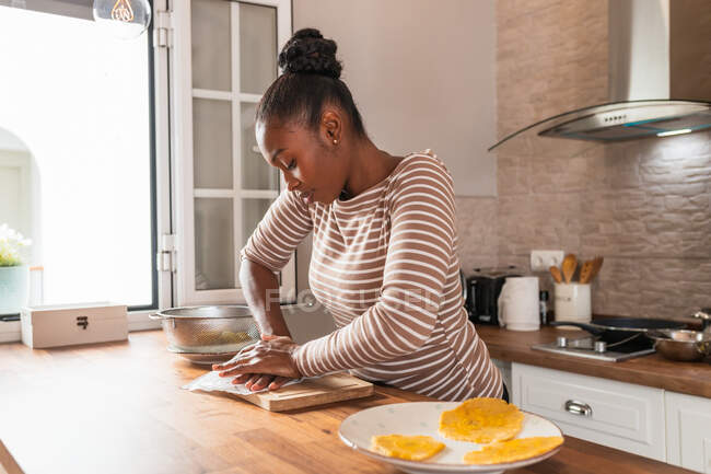 Jeune afro-américaine femme écrasant plantain frais sur planche à découper tout en préparant patacones à la maison — Photo de stock