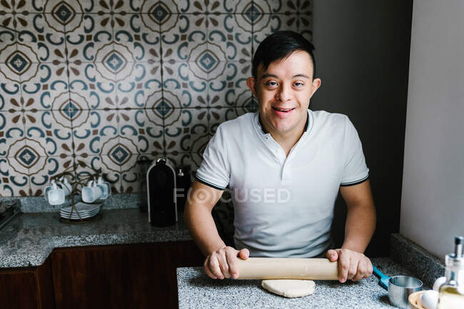 Adolescente étnico positivo com síndrome de Down massa rolante com rolo enquanto cozinha na cozinha e olhando para a câmera — Fotografia de Stock