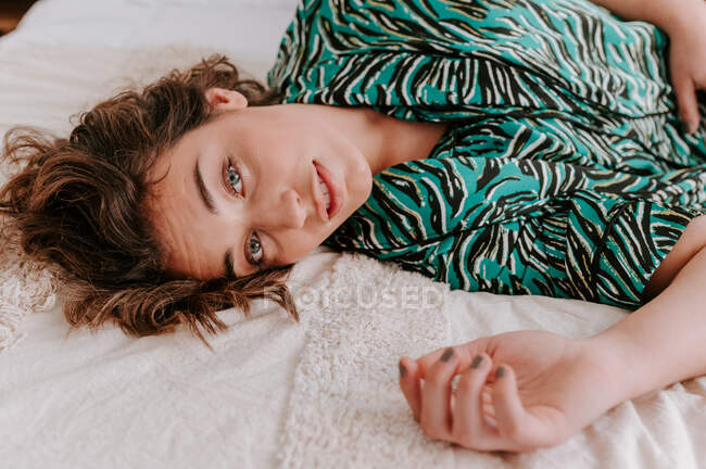 High angle of tranquil female in domestic clothes resting on soft bed at home and looking at camera — Stock Photo