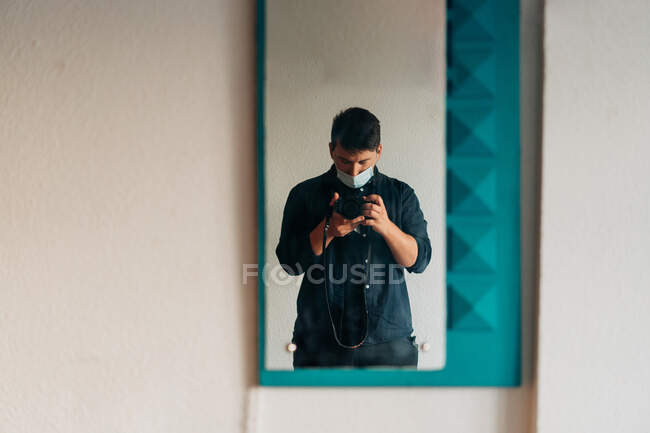 Mirror hanging on wall and reflecting man in casual clothes and mask taking photo — Stock Photo