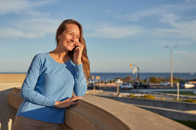 Dame adulte souriante ayant un appel téléphonique tout en s'appuyant sur la clôture près de l'océan dans la rue de la ville dans la journée ensoleillée — Photo de stock