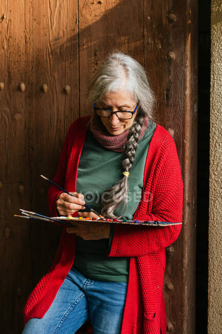 Задоволена старша жінка-художниця з палітрою фарб і пензлями, що стоять біля дерев'яних дверей — стокове фото