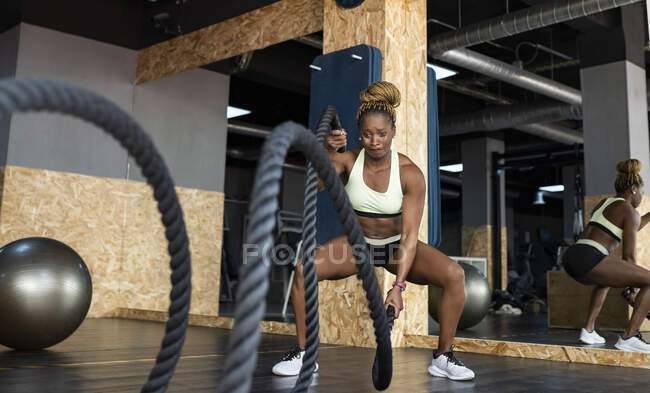 Сильная афро-американская спортсменка в спортивной одежде с боевыми канатами во время интенсивных тренировок в тренажерном зале — стоковое фото