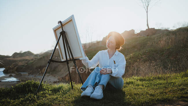 Полное тело молодой женщины в берете и стильной одежде, сидящей на травянистой лужайке возле холмов, рисуя картину кистью на холсте на мольберте в солнечный день под небом — стоковое фото