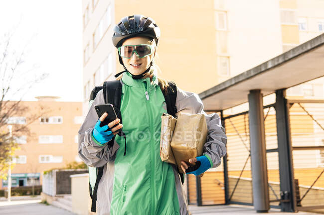 Livraison heureuse femme portant des boîtes enveloppées et naviguant sur la carte GPS sur le téléphone mobile tout en se tenant dans la rue le jour ensoleillé — Photo de stock