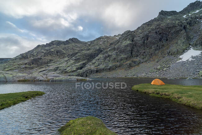 Tenda arancione situato sulla riva erbosa del lago Laguna Grande contro Sierra de Gredos catena montuosa e cielo nuvoloso in Avila, Spagna — Foto stock