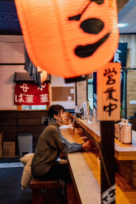 Vista laterale della giovane donna asiatica in abbigliamento casual seduta al bancone in legno in attesa di ordine in ramen bar — Foto stock