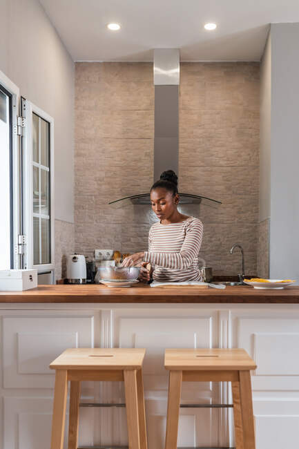 Donna etnica preparare pataconi mentre in piedi a tavola con ciotola e piatto sotto lampade lucide — Foto stock
