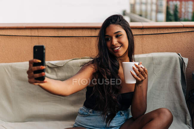Счастливая молодая латиноамериканка с кружкой горячего напитка, улыбающаяся и делающая селфи, сидя на диване на балконе — стоковое фото