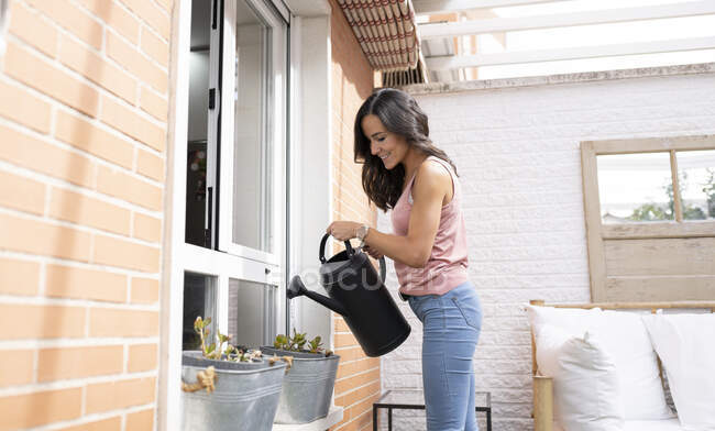 Вид сбоку жизнерадостной женщины, поливающей горшечные цветы на террасе летом — стоковое фото