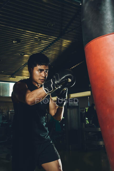 Молодой ориентированный азиат тренирует бокс выполняя удары, тренируясь с тяжелой боксерской грушей в современном тренажерном зале — стоковое фото