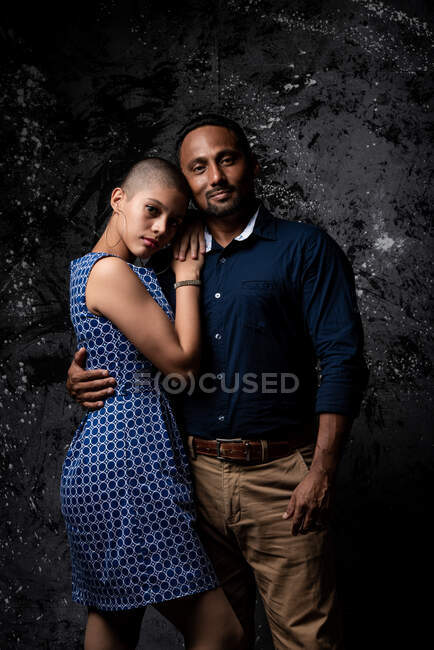 Tender ethnic man embracing woman on dark background in studio looking at camera — Stock Photo