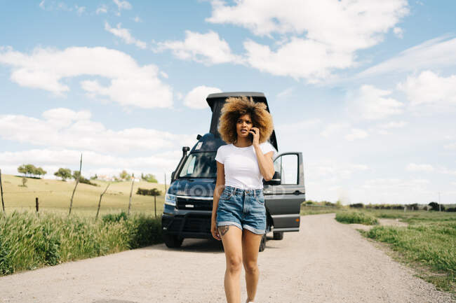 Worried young African American female with curly hair talking on mobile phone while asking for help from repair service after accident with camper van in countryside — Stock Photo