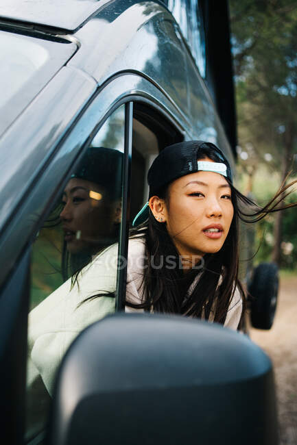 Happy young Asian female traveler in cap looking out open window of camper van and enjoying freedom during trip through summer countryside — Stock Photo