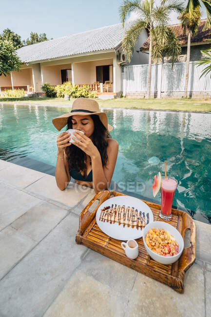 Alegre turista femenina apoyada en la piscina mientras bebe café contra bandeja con delicioso desayuno a la luz del sol - foto de stock