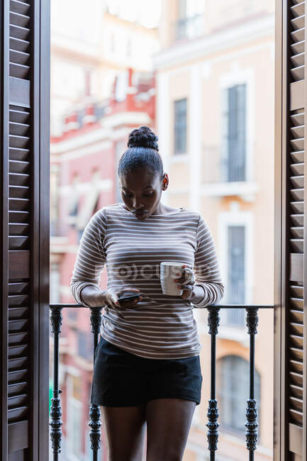 Afroamerikanerin mit Tasse Heißgetränk surft zu Hause auf Handy zwischen Rollläden im Internet — Stockfoto