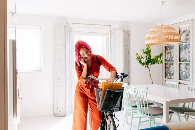 Молода креативна жінка-дизайнерка в модному вбранні та окулярах розмовляє на смартфоні, стоячи з велосипедом в сучасній світлій квартирі — стокове фото