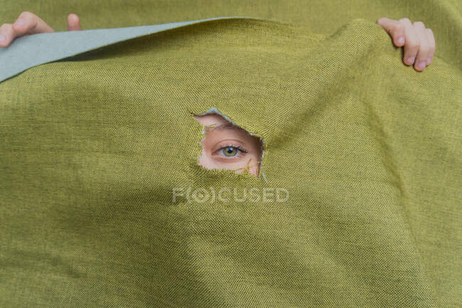 Unerkennbares junges grünäugiges Weibchen guckt durch aufgerissenes Loch in grünem Tuch hinaus — Stockfoto