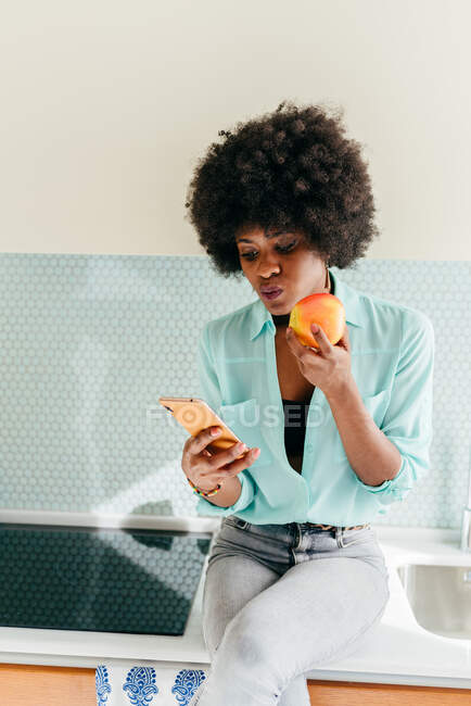 Современная красивая афроамериканка со смартфоном в руке сидит на кухонном столе, смотрит в сторону дома и ест яблоко — стоковое фото