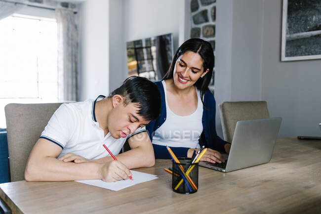 Ethnic teenage boy with Down syndrome drawing with pencils on paper while sitting at table with female freelancer working on laptop at home — Stock Photo